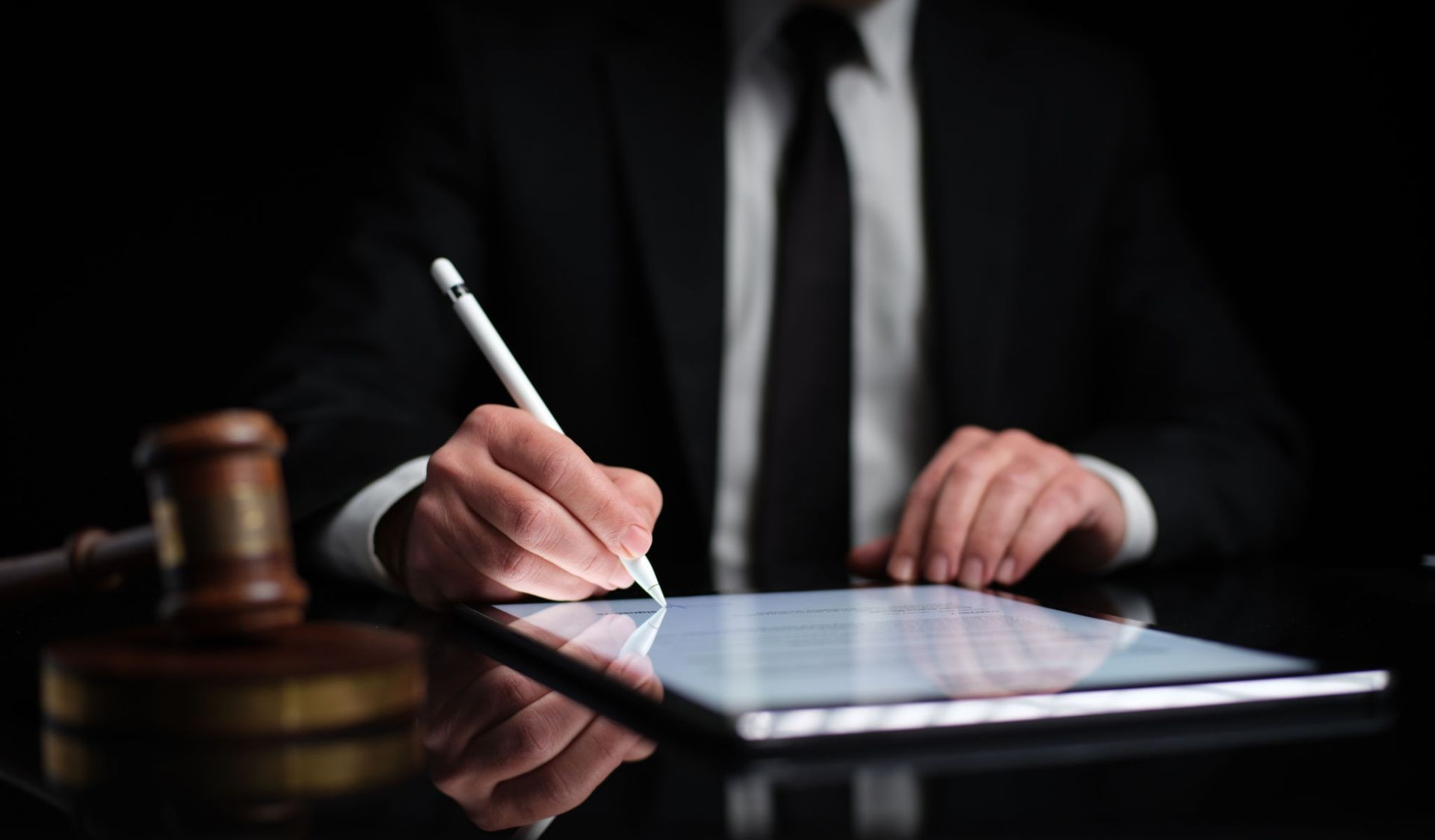Person in suit signing document on digital tablet with stylus, gavel on the table.